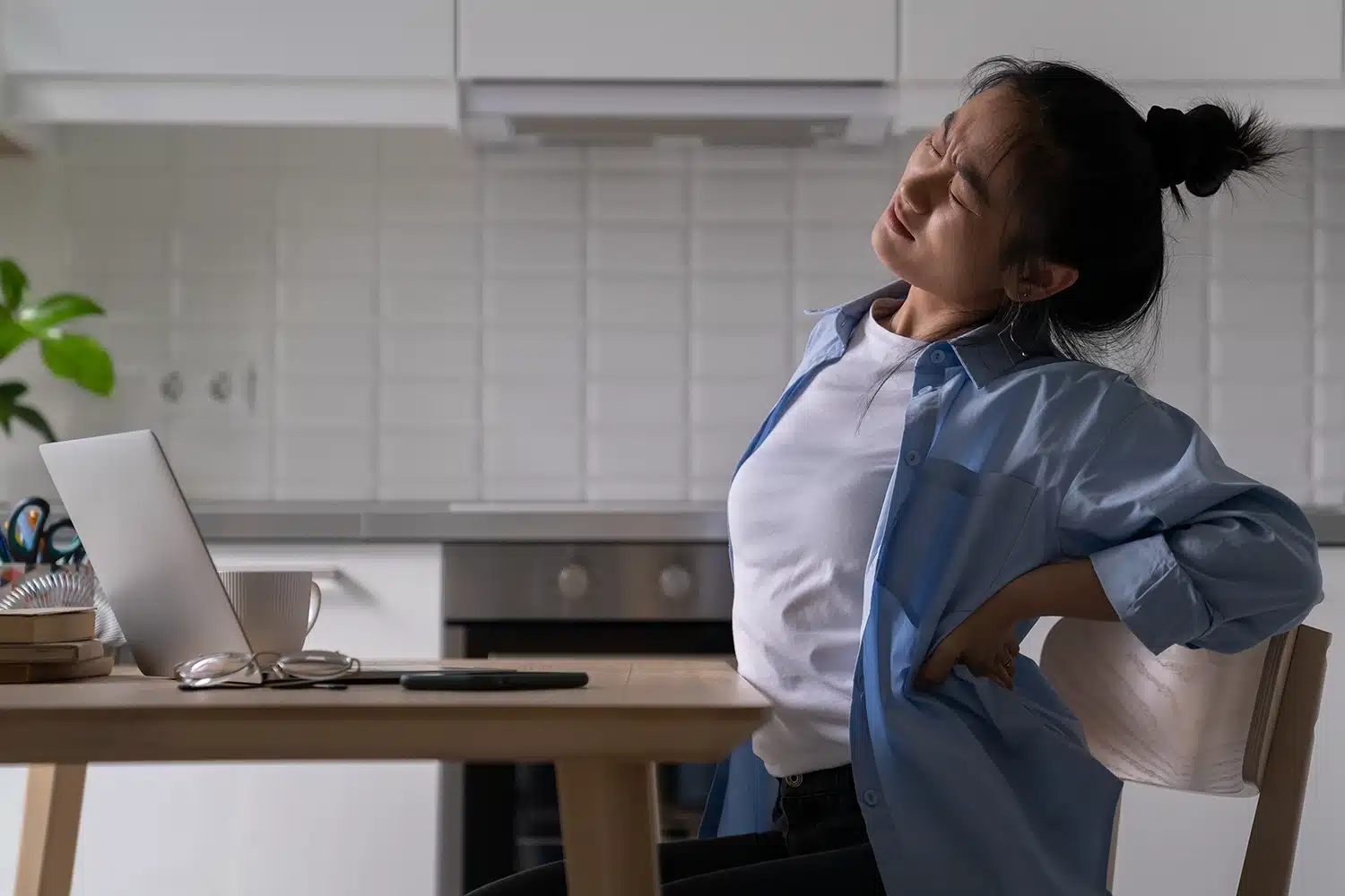 A woman experiencing lower back pain while sitting at a desk with a laptop and a cup. Lower Back Pain When Sitting for Long Hours Causes, Treatment & Relief - Spire Orthopaedic Centre